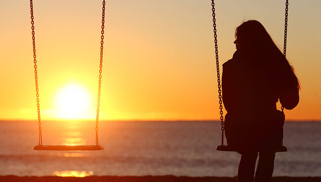 Woman sitting alone next to an empty swing as the sun sets.